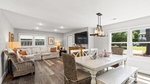 Een moderne woonkamer met een witte bank, rieten stoelen, een eettafel en een televisie.
