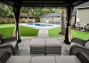 Eine überdachte Terrasse mit Korbmöbeln, einem Steinweg, einem Pool und einer Rasenfläche.