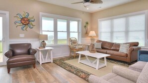 Un salon comprenant un fauteuil en cuir marron, un canapé en cuir marron, une table basse blanche et un fauteuil beige. On y trouve également un ventilateur de plafond, un soleil décoratif accroché au mur et une fenêtre avec des stores.