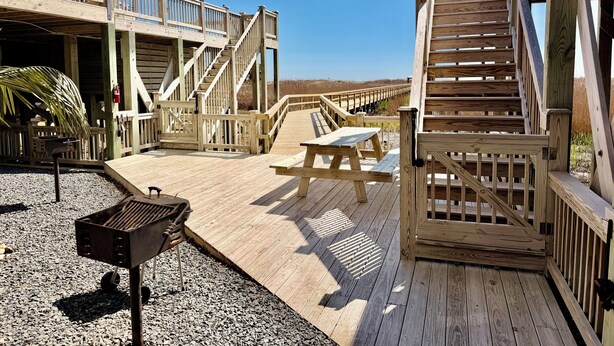 Une terrasse en bois équipée d’un barbecue, de tables de pique-nique et d’un escalier menant à un niveau supérieur.