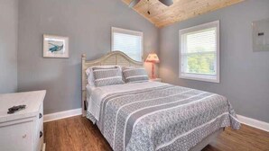 A bedroom with a bed, a ceiling fan, two windows, and a nightstand with a lamp. - Beach Bungalow in Oak Island—a perfect oceanview retreat for up to 8 guests. (Oak Island)