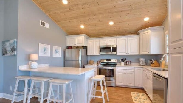 A kitchen with white cabinets, a central island with stools, stainless steel appliances, and a wooden ceiling. - Beach Bungalow in Oak Island—a perfect oceanview retreat for up to 8 guests. (Oak Island)