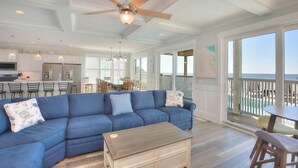 A spacious living room with a blue sectional sofa, a wooden coffee table, and a ceiling fan. - Beach Landing: Spacious 7BR/6BA Oceanfront retreat in Oak Island, sleeps 22 with private pool. (Oak Island)