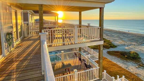 A wooden deck with a pool, a covered area with railings, and a view of the ocean at sunset. - Beach Landing: Spacious 7BR/6BA Oceanfront retreat in Oak Island, sleeps 22 with private pool. (Oak Island)