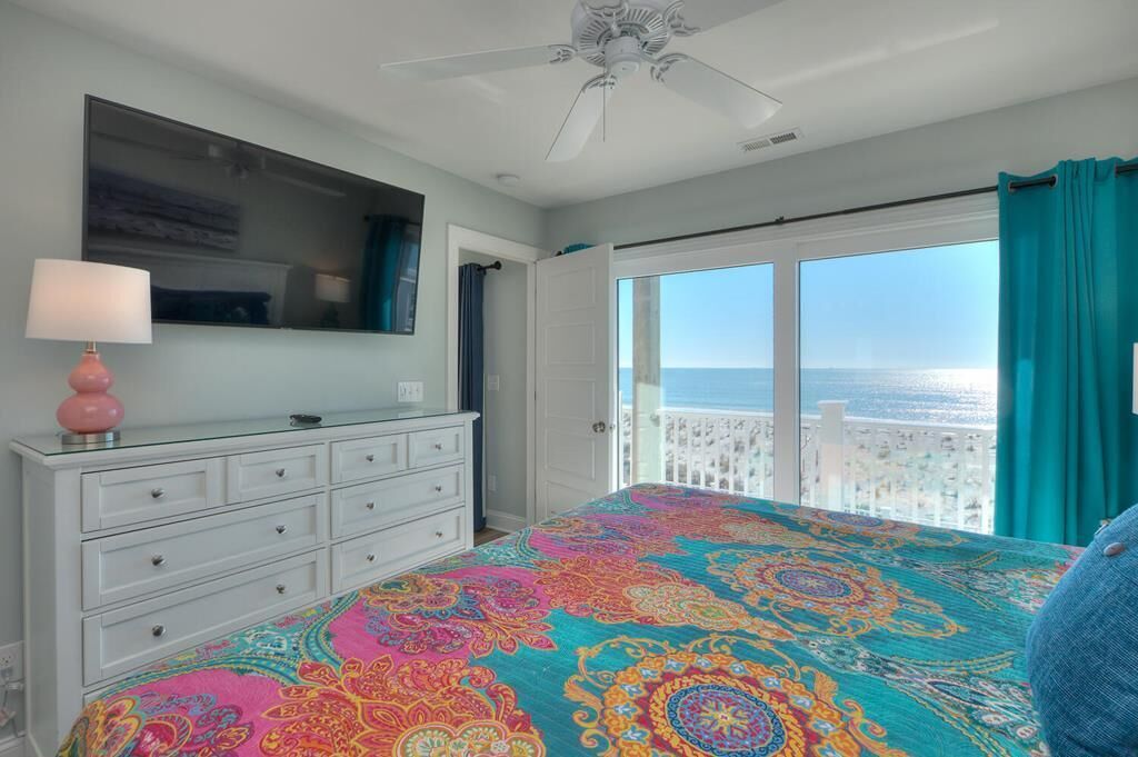 A bedroom with a large bed, a dresser, a ceiling fan, and a view of the ocean through a balcony.