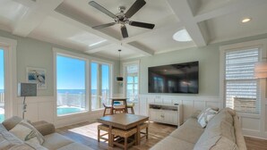 A spacious living room with a large TV, a ceiling fan, a dining table, and a view of the ocean through large windows.