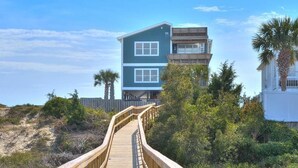 A wooden boardwalk leading to a two-story building with a balcony, surrounded by vegetation and a clear sky. - Bring a Boat! Oceanview/Soundfront Retreat with Gameroom/Pool/HT (Oak Island)