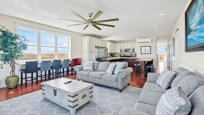 A spacious living room with a grey sofa, a coffee table, and a dining area with chairs. - Bring a Boat! Oceanview/Soundfront Retreat with Gameroom/Pool/HT (Oak Island)