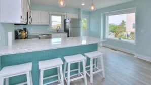 A modern kitchen with white cabinets, a large island with stools, a refrigerator, and pendant lighting. - Beach Tonic: Oceanfront retreat in Oak Island, this 5BR home accommodates up to 12 guests. (Oak Island)