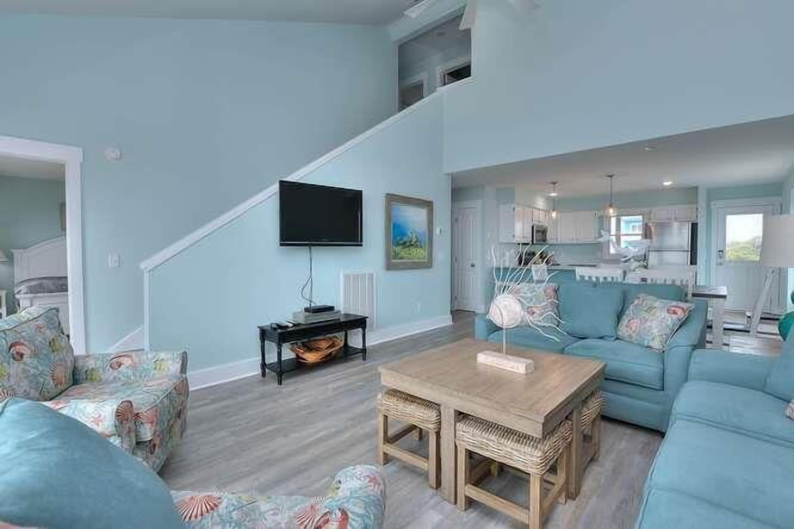 A spacious living room with a staircase, a flat-screen TV, a dining area, and a kitchen in the background.