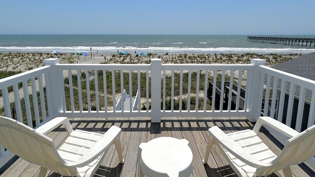 A wooden deck with white railings overlooking a beach with a pier in the distance. - Beach Tonic: Oceanfront retreat in Oak Island, this 5BR home accommodates up to 12 guests. (Oak Island)