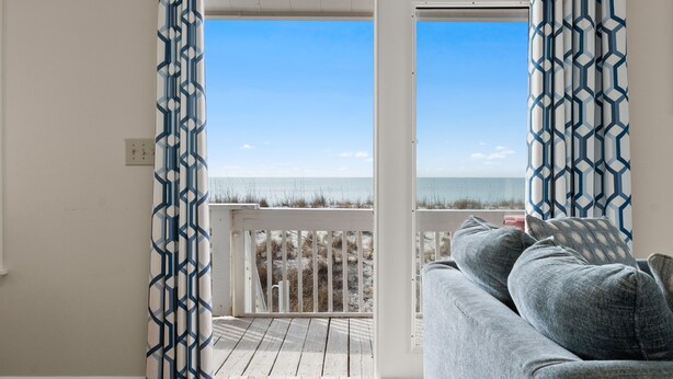 Un balcón con vista a la playa, barandilla blanca y sofá gris con cojines.