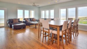 A spacious living room with a wooden dining table and chairs, a sofa set, and a large window offering a view of the ocean. - Bright Horizons: Experience oceanfront luxury in this 7-bedroom, 6-bath retreat in Oak Island! (Oak Island)