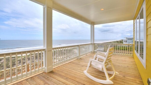 A covered porch with a wooden deck, white rocking chairs, and a view of the ocean. - Bright Horizons: Experience oceanfront luxury in this 7-bedroom, 6-bath retreat in Oak Island! (Oak Island)