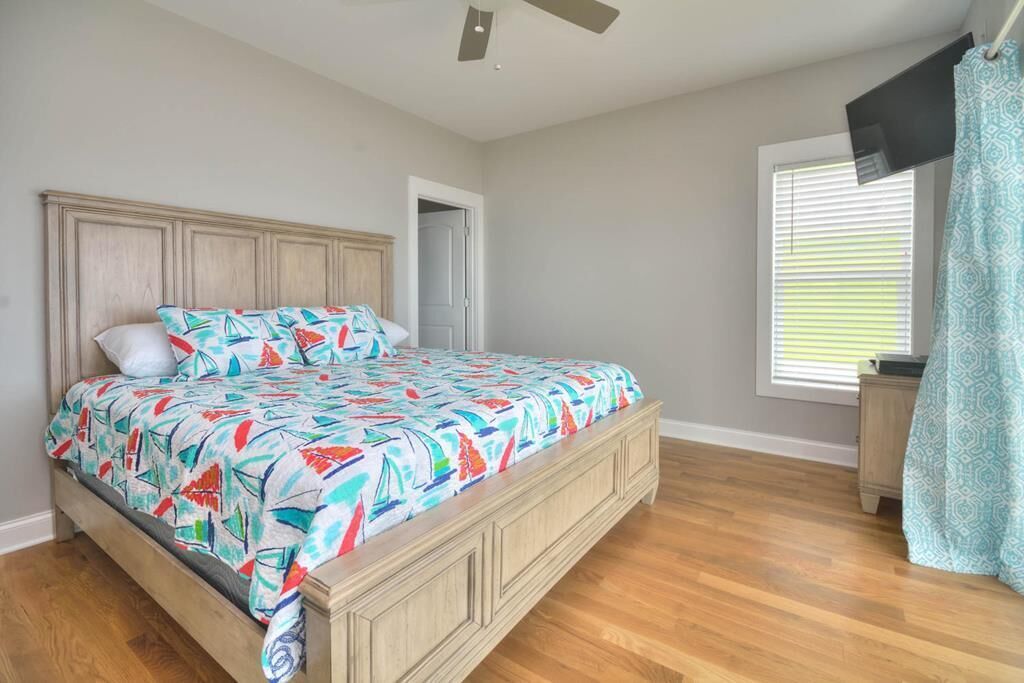 A bedroom with a wooden bed, a ceiling fan, a window with blinds, and a TV mounted on the wall.