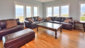 A spacious living room with leather sofas, a glass coffee table, and hardwood flooring. Large windows offer a view of the ocean. - Bright Horizons: Experience oceanfront luxury in this 7-bedroom, 6-bath retreat in Oak Island! (Oak Island)