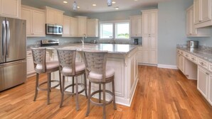 A kitchen with white cabinets, a stainless steel refrigerator, a microwave, and a large island with bar stools. - Bright Horizons: Experience oceanfront luxury in this 7-bedroom, 6-bath retreat in Oak Island! (Oak Island)