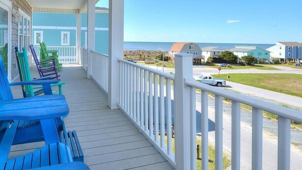 Une terrasse en bois agrémentée de chaises Adirondack aux couleurs vives, d’une balustrade blanche et offrant une vue sur l’océan et les maisons qui se dessinent à l’arrière-plan.