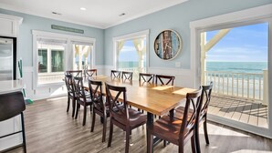 A dining area with a wooden table and chairs, large windows with a view of the ocean, and a clock on the wall. - Experience oceanfront luxury at Chasing the Sun, a spacious 7-bedroom retreat in Oak Island. (Oak Island)