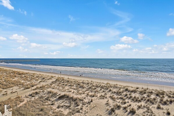 A beach with a wooden pier extending into the ocean, sandy dunes, and a clear sky.
