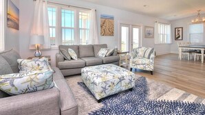 A spacious living room with a grey sofa, matching ottoman, and armchairs, a dining area with a white table, and large windows allowing natural light. - Comin in Hot: Spacious 4-bedroom oceanview home in Oak Island with private pool! (Oak Island)