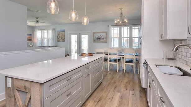 A modern kitchen with white cabinetry, a large island, pendant lighting, and a dining area with chairs and a table. - Comin in Hot: Spacious 4-bedroom oceanview home in Oak Island with private pool! (Oak Island)