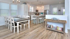 A modern kitchen with white cabinetry, a central island, bar stools, and a bookshelf. - Comin in Hot: Spacious 4-bedroom oceanview home in Oak Island with private pool! (Oak Island)