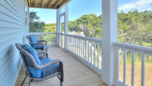 A covered porch with two wicker chairs, a white railing, and a view of trees. - Comin in Hot: Spacious 4-bedroom oceanview home in Oak Island with private pool! (Oak Island)