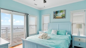 A bedroom with a bed, a nightstand, a lamp, and a painting. There is a view of the beach through the window. - Experience oceanfront luxury at Cordoba, a spacious 7-bedroom retreat in Oak Island! (Oak Island)