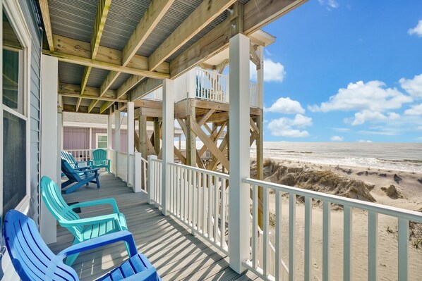 Een houten terras met blauwe stoelen en een leuning met uitzicht op een strand.