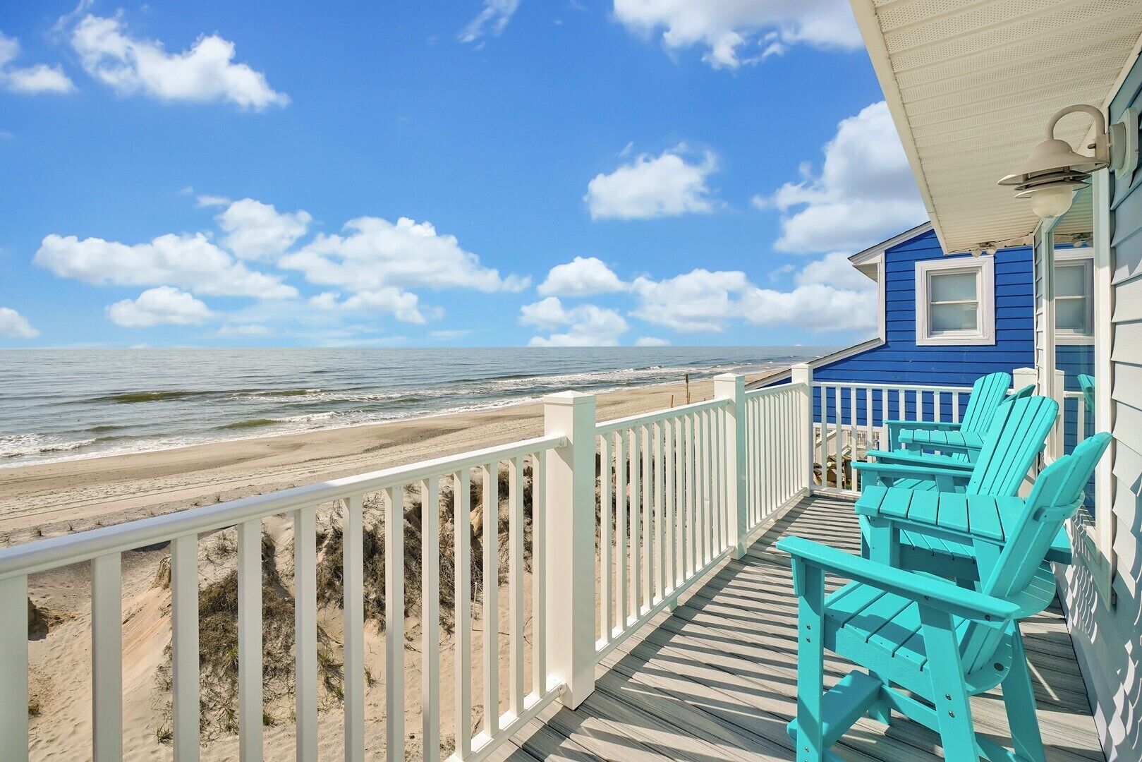 Een strandhuis met een houten terras, turquoise Adirondack-stoelen en uitzicht op de oceaan.