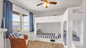 Une chambre avec un lit superposé, une chaise en osier, un ventilateur de plafond et une vue sur l’océan.