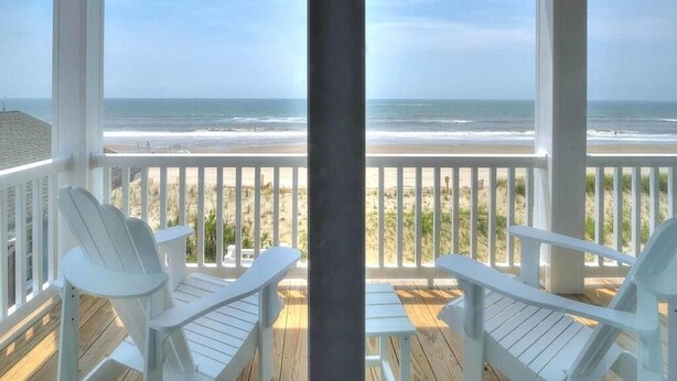Terraza de madera con sillas blancas con vista a una playa.
