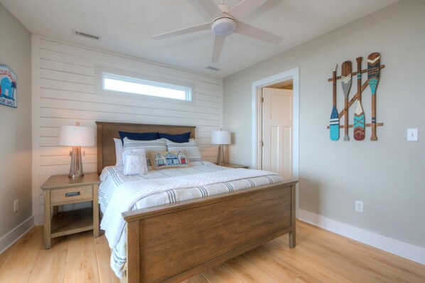A bedroom with a wooden bed, two bedside tables with lamps, a ceiling fan, a window, and a wall with paddleboard decorations. - Experience beachfront luxury at Fishizzle in Oak Island, perfect for family getaways. (Oak Island)