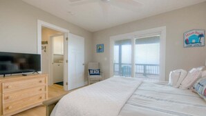 Un dormitorio con cama, televisor sobre un aparador, silla con almohada de playa, balcón con vistas y un letrero que dice 'BEACH'.