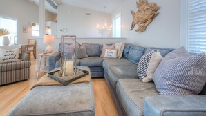 Una sala de estar con un sofá gris, un sillón estampado, una mesa de vidrio con una vela y una obra de arte de madera en la pared.