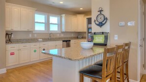 A kitchen with white cabinets, a granite countertop, and a large island with a bowl. There is a clock on the wall and a window above the sink. - Discover Gypsea, a spacious 4-bedroom oceanview home in Oak Island, perfect for 10 guests! (Oak Island)