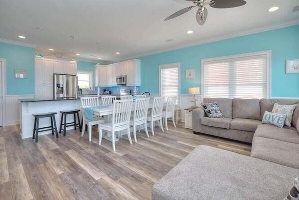 A modern kitchen with white cabinets, a dining area with white chairs, and a beige sofa with a window view.