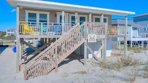 Un edificio frente a la playa con una escalera de madera que lleva a un balcón, un letrero que anuncia una playa mejor, y un cielo azul despejado.
