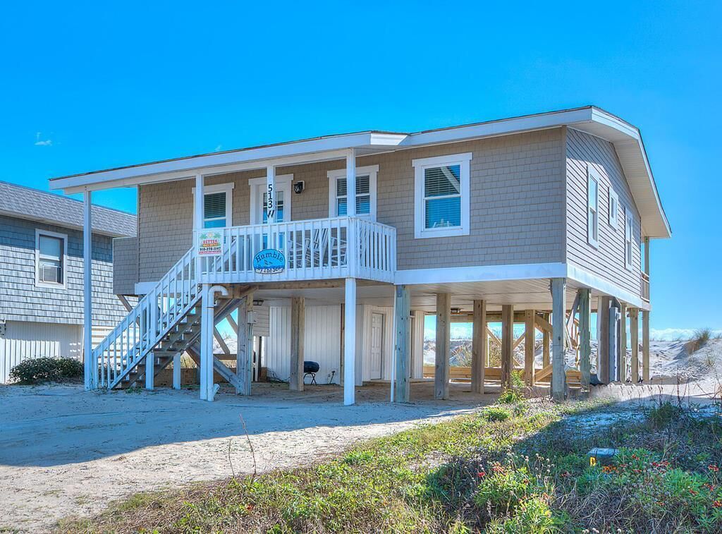 A two-story beach house with a balcony, a staircase, and a sandy beach in the background.