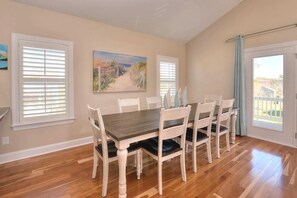 A dining area with a wooden table and white chairs, a painting on the wall, and a view through a door.