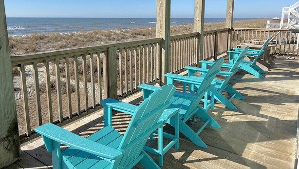 Um deck de madeira com cadeiras Adirondack turquesa voltadas para a praia.