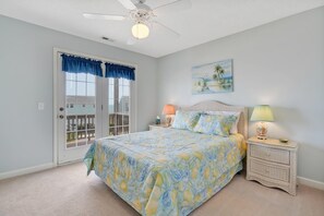 Ein Schlafzimmer mit einem Bett, zwei Nachttischen, einem Deckenventilator, einem Fenster mit blauen Vorhängen und einem Bild an der Wand.