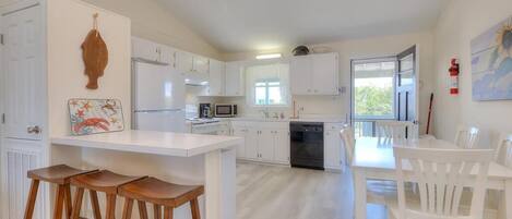 Una cocina moderna con armarios blancos, una isla central con taburetes y un comedor con sillas blancas.
