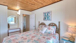 A bedroom with a wooden ceiling, a bed with floral bedding, a bedside table with a lamp, a mirror, and a door leading to another room.
