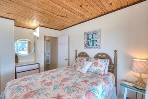 A bedroom with a wooden ceiling, a bed with floral bedding, a bedside table with a lamp, a mirror, and a door leading to another room.