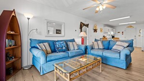 A living room with two blue sofas, a glass coffee table, a ceiling fan, and a bookshelf.