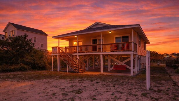 A two-story beach house with a balcony, a staircase, and a sandy beach in the foreground.