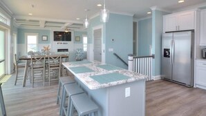 A kitchen with a central island, bar stools, a refrigerator, and a dining area with chairs and a table. - Experience luxury in this oceanfront 5-bedroom retreat in Oak Island. (Oak Island)