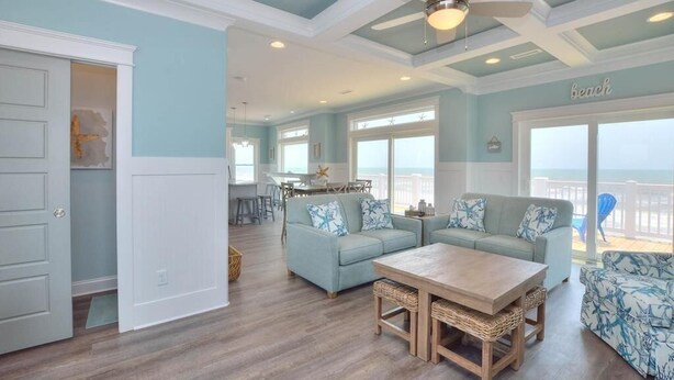 A spacious living room with a large window, a ceiling fan, and a view of the beach. - Experience luxury in this oceanfront 5-bedroom retreat in Oak Island. (Oak Island)
